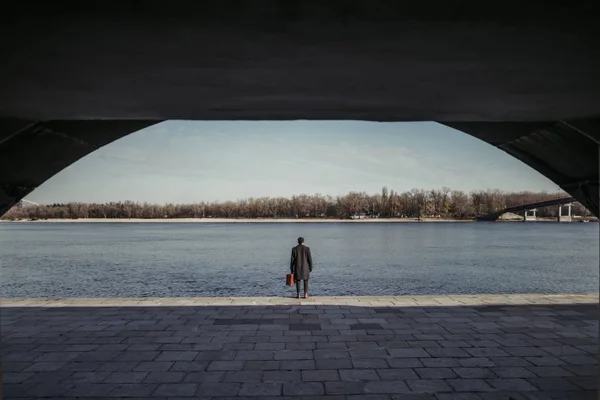 Bel homme adulte avec valise debout sur la rive de la rivière — Photo de stock