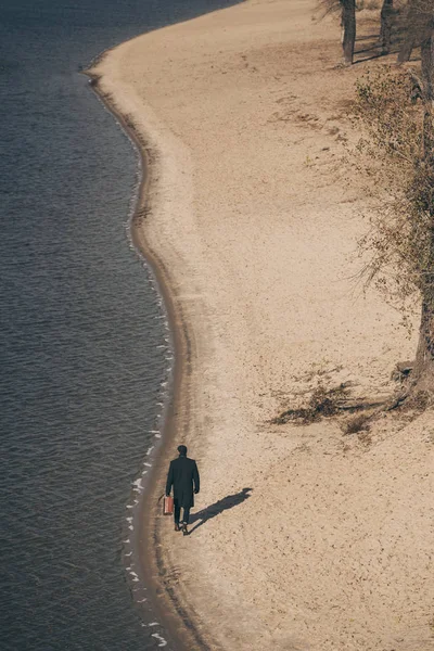 Vista de ángulo alto del hombre en abrigo con maleta caminando por la playa de arena en otoño - foto de stock