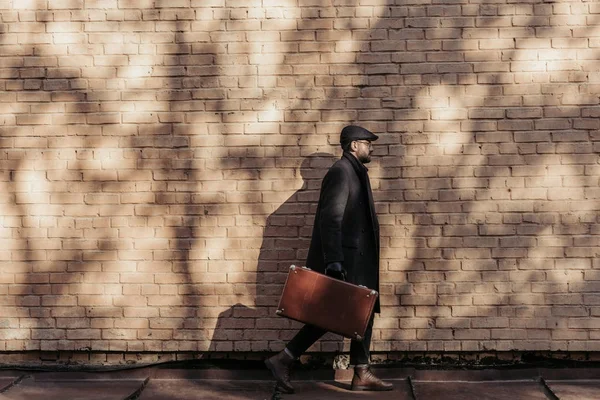 Vista lateral do homem adulto com mala andando na frente da parede de tijolo — Fotografia de Stock