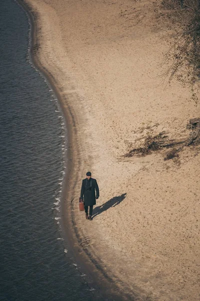 Vista de ángulo alto del hombre con el equipaje caminando por la playa de arena en otoño - foto de stock