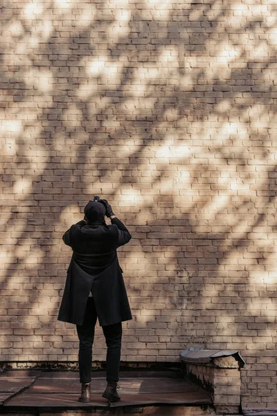 Back view of stylish man with camera taking photo of brrick wall with tree shadows — Stock Photo