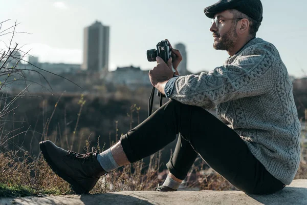 Homem adulto bonito com câmera de filme vintage sentado na estrada rural no dia ensolarado e olhando para longe — Fotografia de Stock