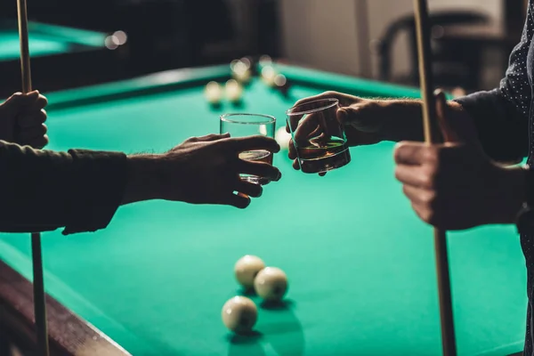 Cropped image of male hands with cues and glasses with alcohol infront of billiard table at bar — Stock Photo