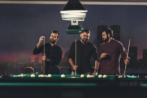 Compañía de hombres guapos exitosos de pie junto a la mesa de billar con bebida en el bar - foto de stock