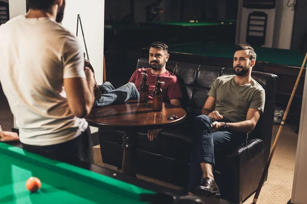 Hombres caucásicos jóvenes sentados en el sofá y bebiendo cerveza junto a la mesa de billar en el bar - foto de stock