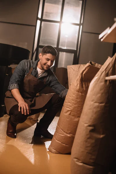 Sorrindo torrador de café jovem olhando para sacos de papel enquanto trabalhava em café — Fotografia de Stock