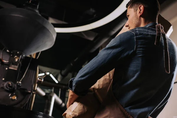 Visão traseira do torrador de café segurando saco de papel em mãos enquanto em pé na máquina de torrefação — Fotografia de Stock