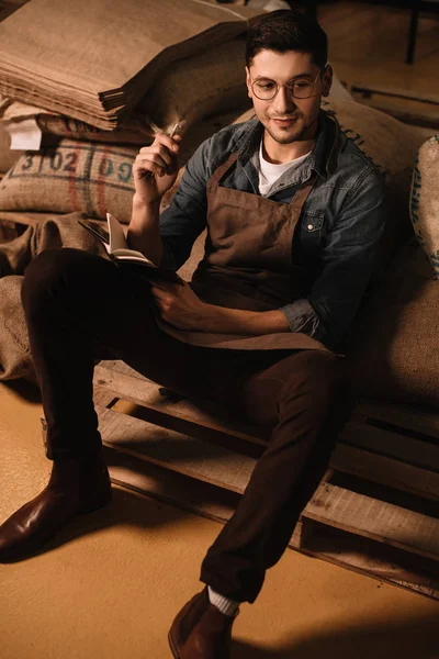 Pensive coffee roaster in eyeglasses and apron looking away while making notes in notebook — Stock Photo