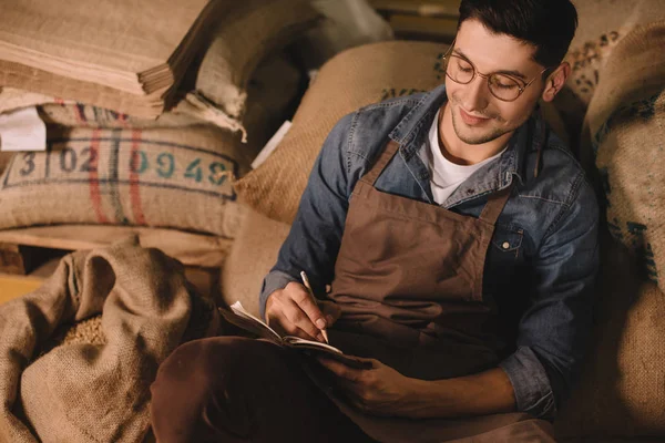 Portrait of smiling coffee roaster in eyeglasses and apron making notes in notebook — Stock Photo