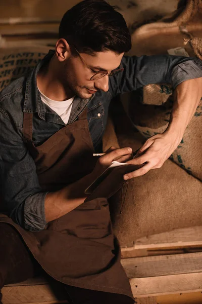 Retrato de tostador de café en anteojos y delantal haciendo notas en cuaderno - foto de stock