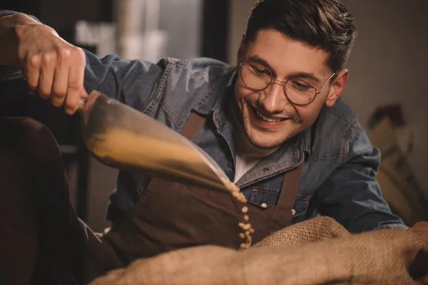 Retrato de trabajador sonriente en delantal vertiendo granos de café en saco - foto de stock