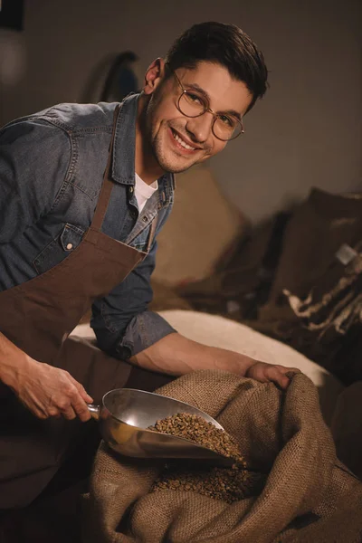 Sonriente trabajador en delantal verter granos de café en saco bolsa - foto de stock