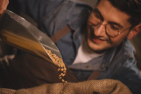 Selective focus of smiling worker in apron pouring coffee beans into sack bag — Stock Photo