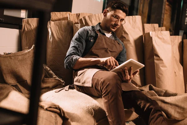 Joven trabajador en delantal usando tableta mientras está sentado en saco - foto de stock