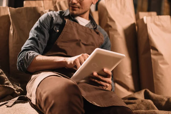 Cropped shot of worker in apron with tablet sitting on sack bag — Stock Photo