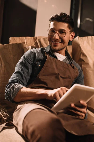 Retrato del trabajador sonriente con la tableta mirando a la cámara mientras está sentado en el saco - foto de stock