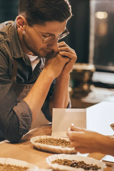 Side view of pensive coffee shop worker looking at coffee beans in bowl on wooden tabletop — Stock Photo