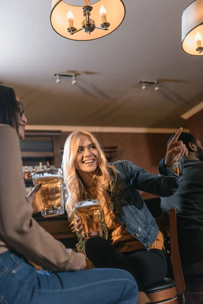 Two young women drinking beer at bar and pointing on something — Stock Photo