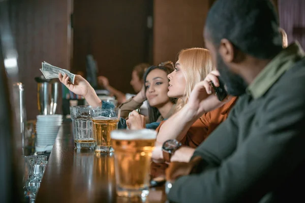 Hermosa chica con dinero pagando por la bebida en el bar con amigos - foto de stock