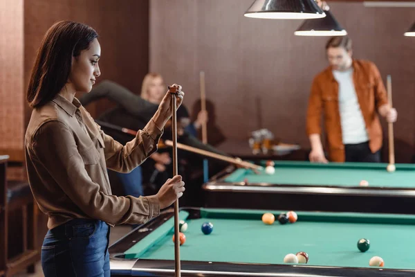 Joven chica tiza señal al lado de la mesa de billar en el bar con amigos — Stock Photo