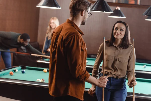 Молодой человек и женщина стоят у бильярдного стола и смотрят друг на друга в баре с друзьями — стоковое фото