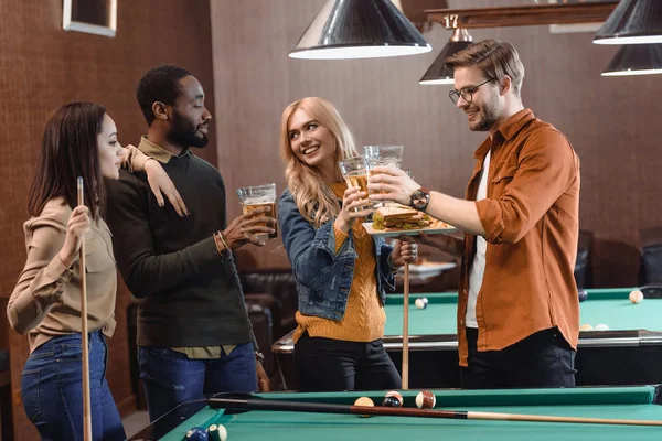 Company of multiethnic friends eating and drinking beside pool table at bar — Stock Photo