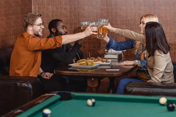 Compañía de amigos multiétnicos comiendo y bebiendo al lado de la mesa de billar en el bar - foto de stock