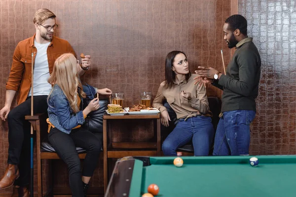 Compagnia di amici multietnici che mangiano e bevono accanto al tavolo da biliardo al bar — Foto stock