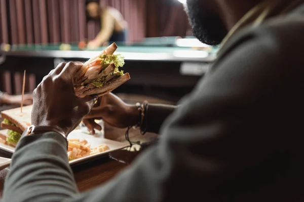 Imagen recortada de hombre amerciano africano comiendo sándwich en el bar - foto de stock