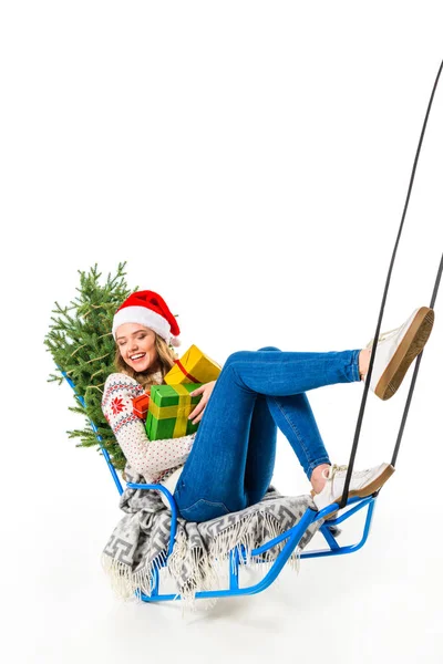 Смеющаяся девушка в шапке Санты сидит на санях с подарками и рождественской ёлкой, изолированная на белом — стоковое фото