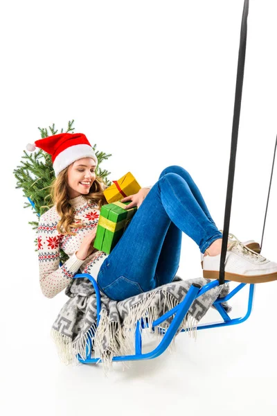 Fröhliche Frau mit Weihnachtsmütze auf Schlitten sitzend mit Geschenkschachteln und Weihnachtsbaum, isoliert auf weiß — Stockfoto