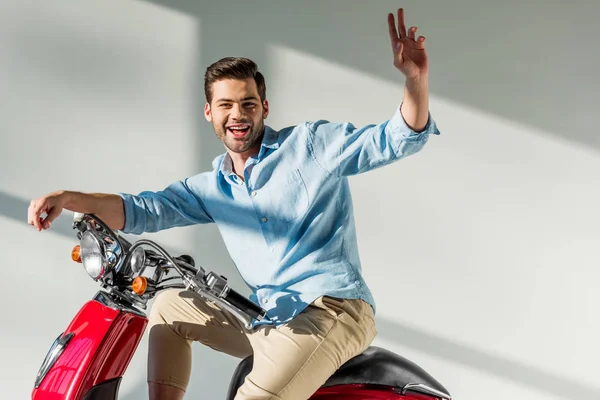 Side view of young man waving to someone while sitting on red scooter — Stock Photo