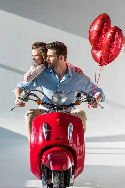 Smiling young couple with red heart shaped balloons riding scooter — Stock Photo