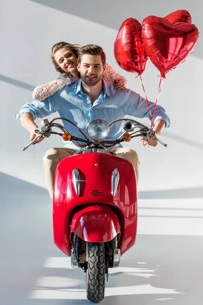 Sorridente giovane coppia con palloncini a forma di cuore rosso equitazione scooter — Foto stock