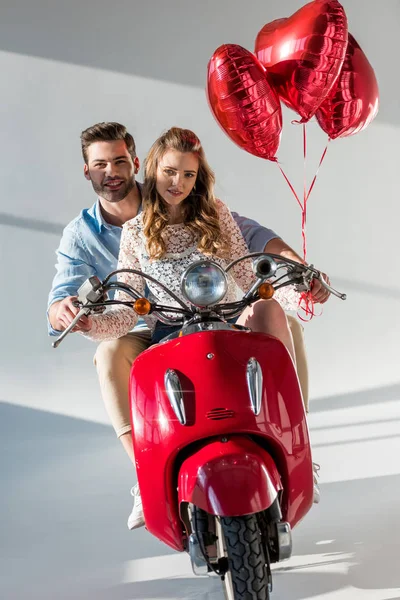 Casal com balões em forma de coração vermelho montando scooter vermelho juntos — Fotografia de Stock