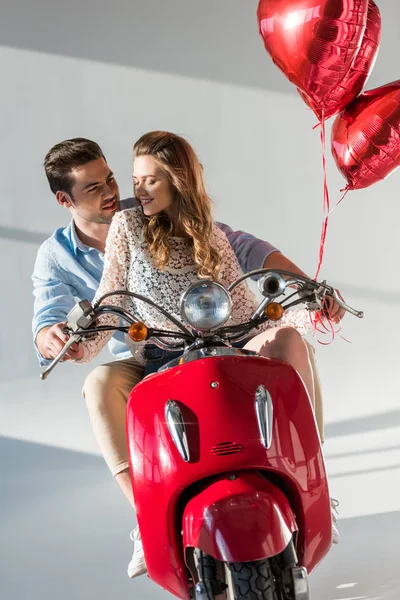 Coppia con palloncini rossi a forma di cuore cavalcando scooter rosso insieme — Foto stock