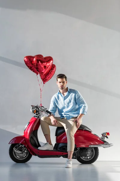 Jeune homme avec des ballons en forme de coeur assis sur scooter rouge — Photo de stock