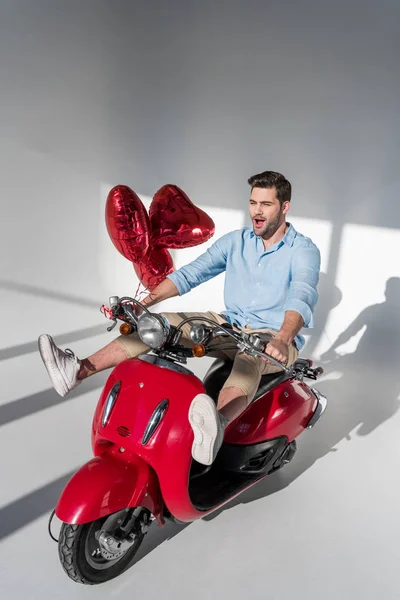 Jeune homme avec des ballons en forme de coeur équitation scooter rouge — Photo de stock