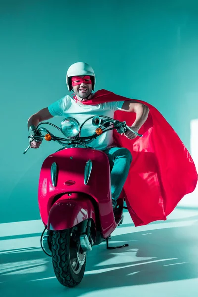 Hombre sonriente en casco protector, máscara de superhéroe y capa de montar scooter rojo - foto de stock