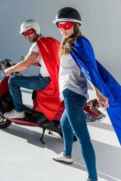 Pareja con estilo en cascos y trajes de superhéroe con scooter rojo mirando a la cámara - foto de stock