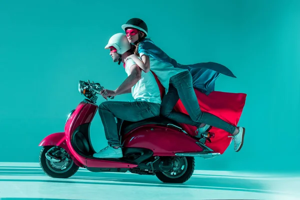 Vista lateral de pareja en trajes de superhéroe montando scooter rojo juntos - foto de stock