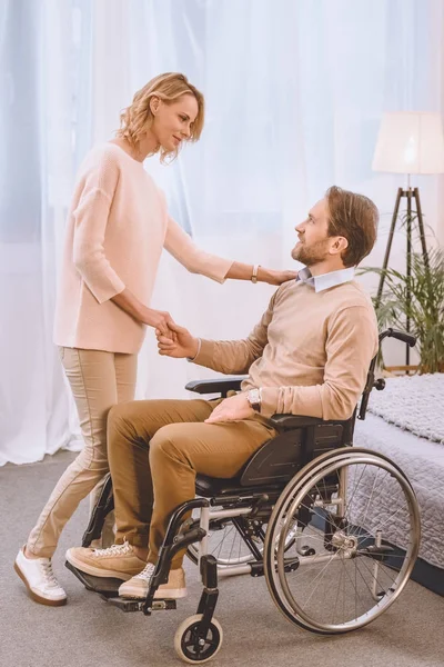 Feliz marido en silla de ruedas y esposa de la mano en el dormitorio - foto de stock