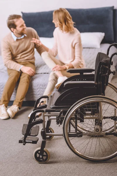 Husband with disability and wife sitting and talking on bed with wheelchair on foreground — Stock Photo