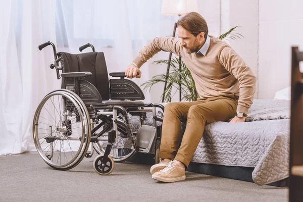 Hombre con discapacidad tratando de sentarse en silla de ruedas desde la cama - foto de stock