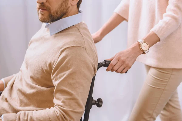 Imagen recortada de la esposa llevando marido en silla de ruedas - foto de stock