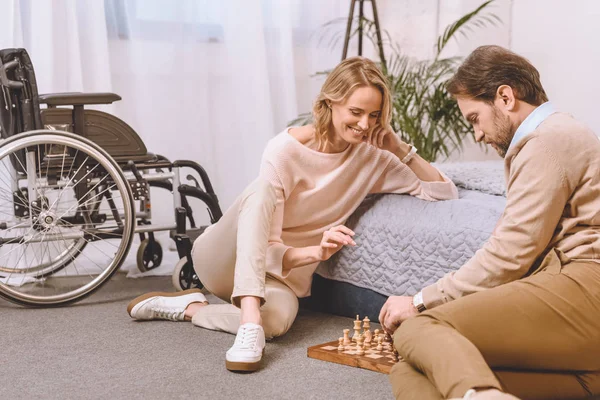 Marido con discapacidad y esposa sonriente jugando ajedrez en el dormitorio - foto de stock