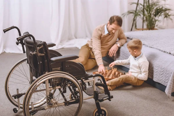 Батько з інвалідністю і син грає в шахи на підлозі з інвалідним візком на передньому плані — стокове фото
