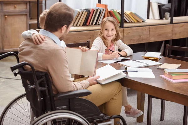 Vater im Rollstuhl unterrichtet lächelnde Kinder zu Hause und umarmt Sohn — Stockfoto