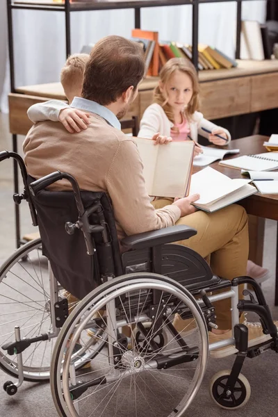 Отец на инвалидной коляске обучает детей дома и обнимает сына — стоковое фото