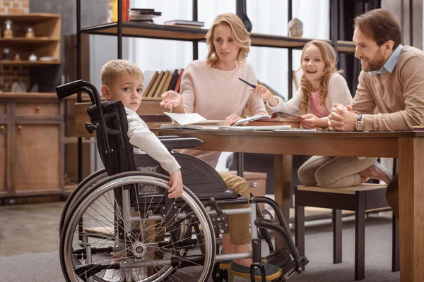 Trauriger Junge im Rollstuhl blickt in die Kamera und seine Verwandten lachen — Stockfoto